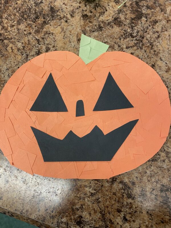 This is a orange pumpkin, it is made out of small pieces of construction paper making layers of orange. There is a green steam and black cut out shapes for the face.
