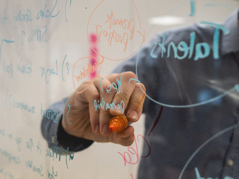 A photo of a clear board that has a ton of words and ideas and is being written on by someone